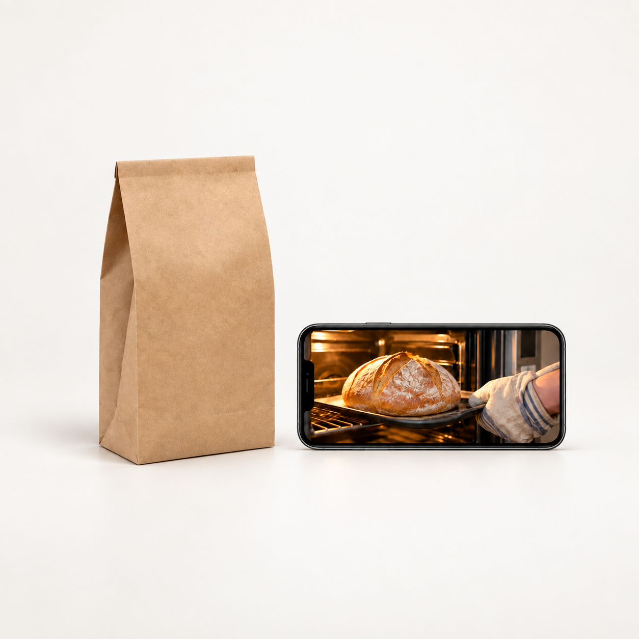 Kaufszenario: Backmischung in einer Papiertüte neben einem Smartphone, auf dessen Display ein Brot aus dem Backofen zu sehen ist